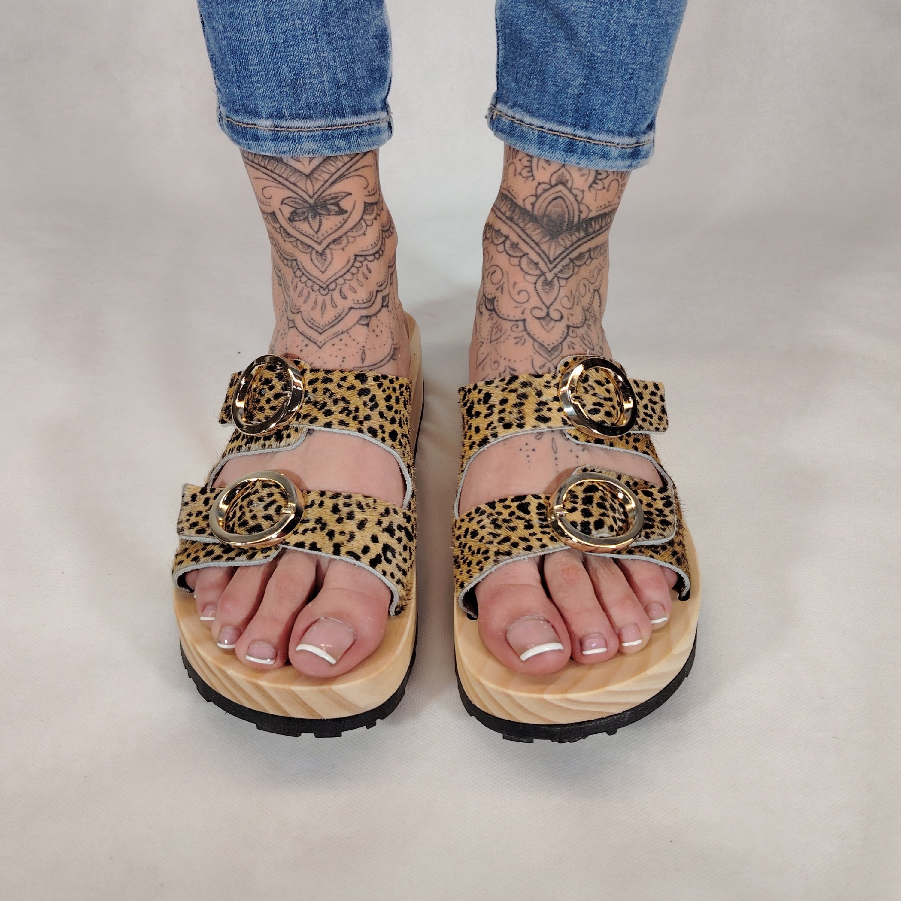 Sandalias tipo zueco de la marca Artichoke con base de madera, dos correas ajustables de cuero y piel sintética de estampado animal print leopard , y hebillas redondas de metal dorado.