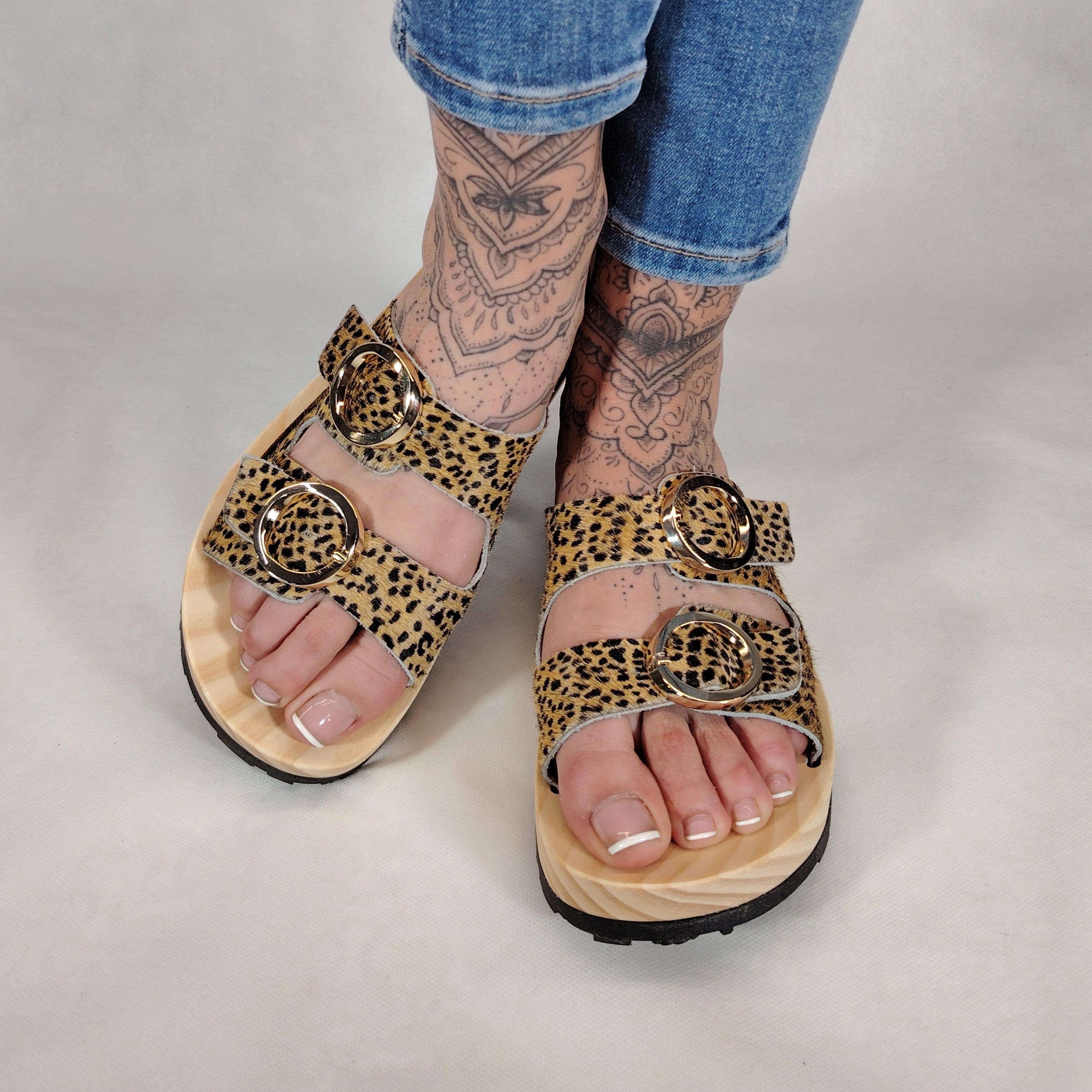 Sandalias tipo zueco de la marca Artichoke con base de madera, dos correas ajustables de cuero y piel sintética de estampado animal print de vaca, y hebillas redondas de metal dorado.