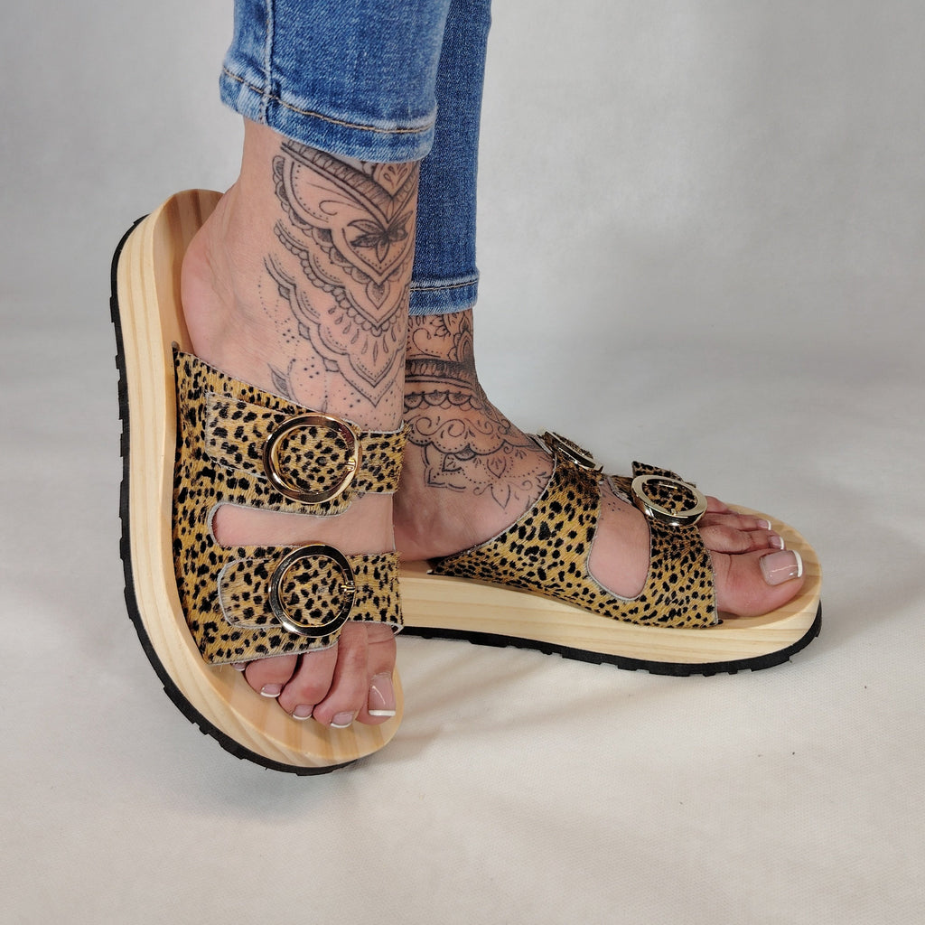 Sandalias tipo zueco de la marca Artichoke con base de madera, dos correas ajustables de cuero y piel sintética de estampado animal print de vaca, y hebillas redondas de metal dorado.