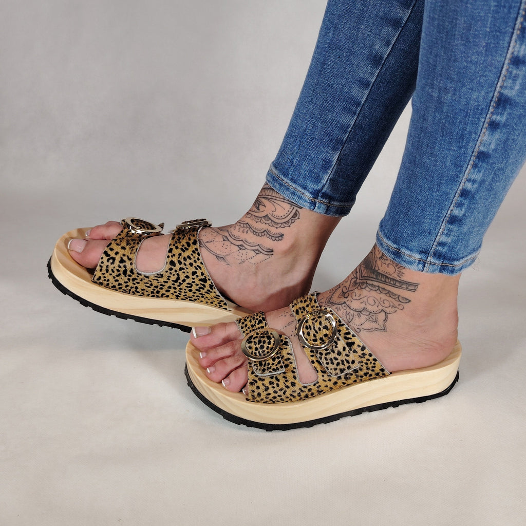 Sandalias tipo zueco de la marca Artichoke con base de madera, dos correas ajustables de cuero y piel sintética de estampado animal print de vaca, y hebillas redondas de metal dorado.