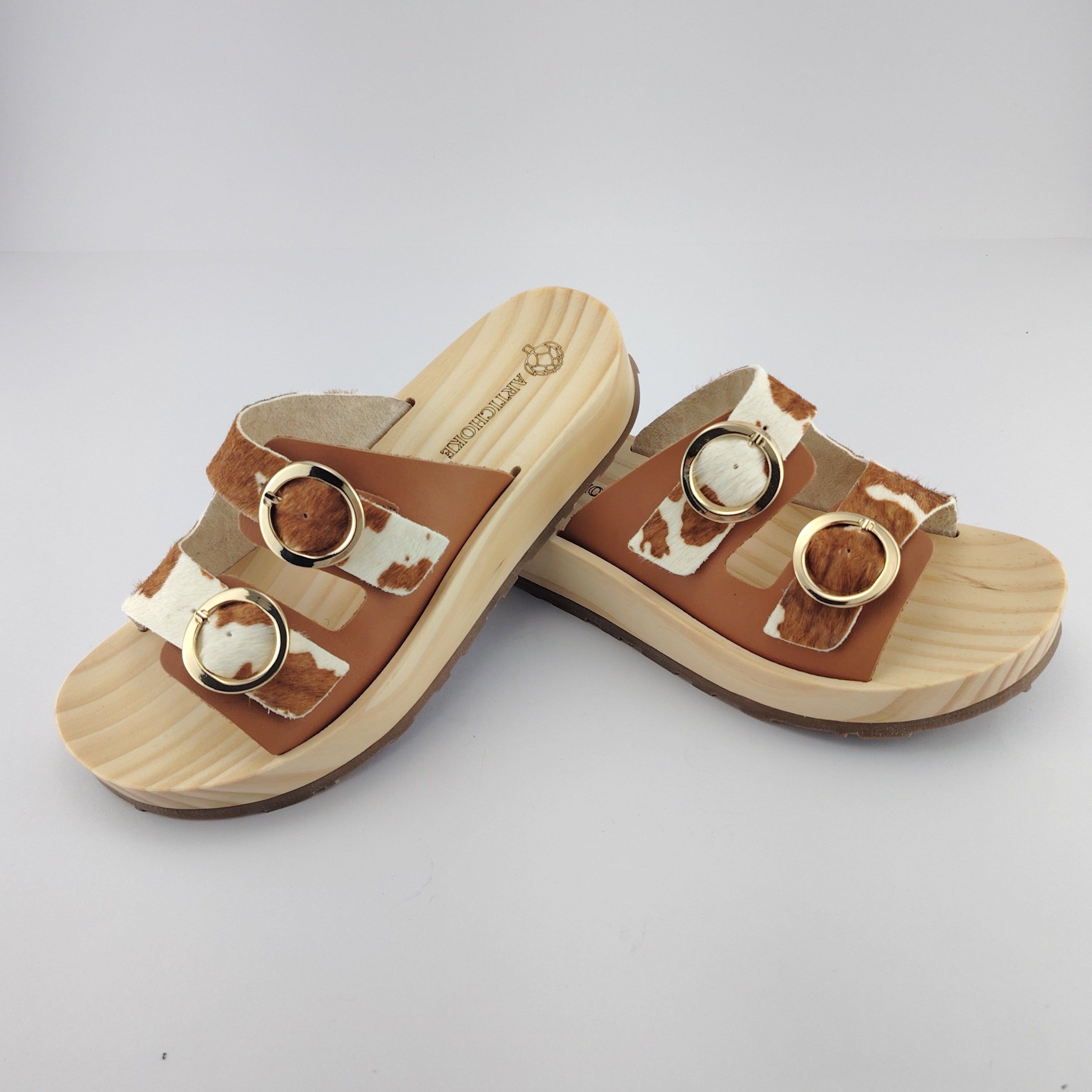 Sandalias tipo zueco de la marca Artichoke con base de madera, dos correas ajustables de cuero y piel sintética de estampado animal print de vaca, y hebillas redondas de metal dorado.