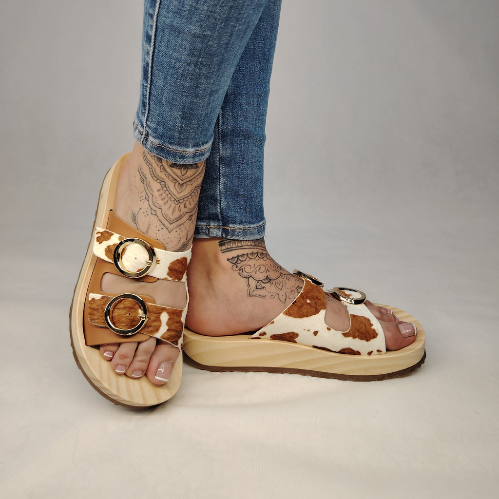Sandalias tipo zueco de la marca Artichoke con base de madera, dos correas ajustables de cuero y piel sintética de estampado animal print de vaca, y hebillas redondas de metal dorado.