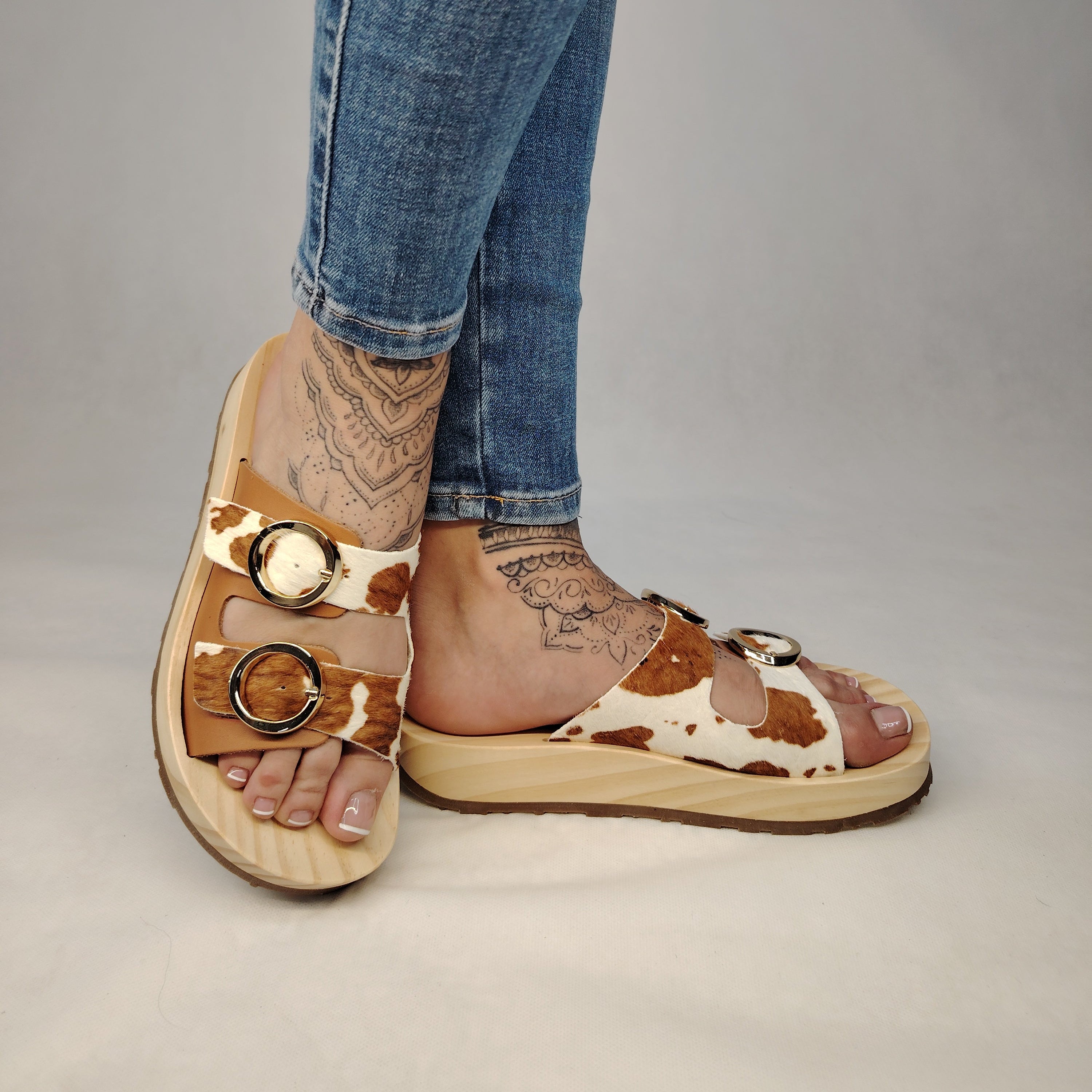 Sandalias tipo zueco de la marca Artichoke con base de madera, dos correas ajustables de cuero y piel sintética de estampado animal print de vaca, y hebillas redondas de metal dorado.