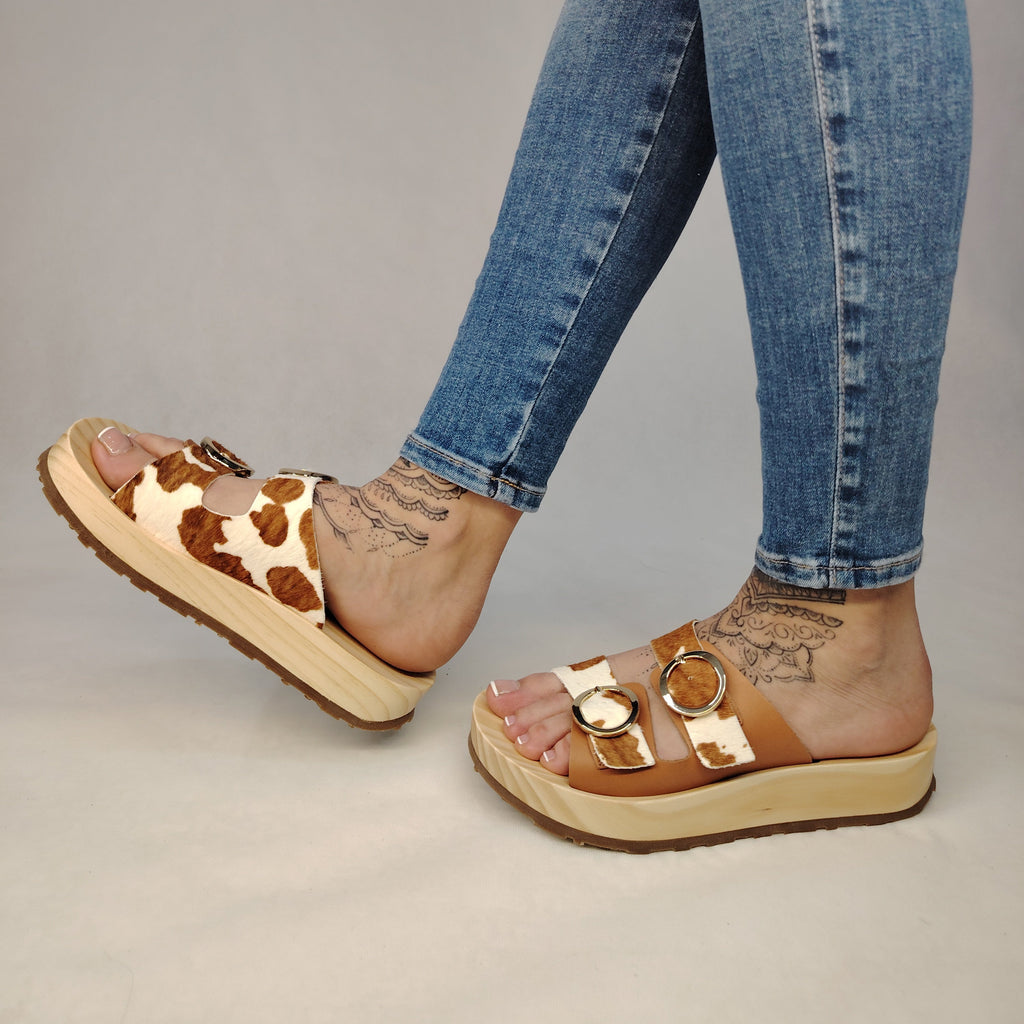 Sandalias tipo zueco de la marca Artichoke con base de madera, dos correas ajustables de cuero y piel sintética de estampado animal print de vaca, y hebillas redondas de metal dorado.
