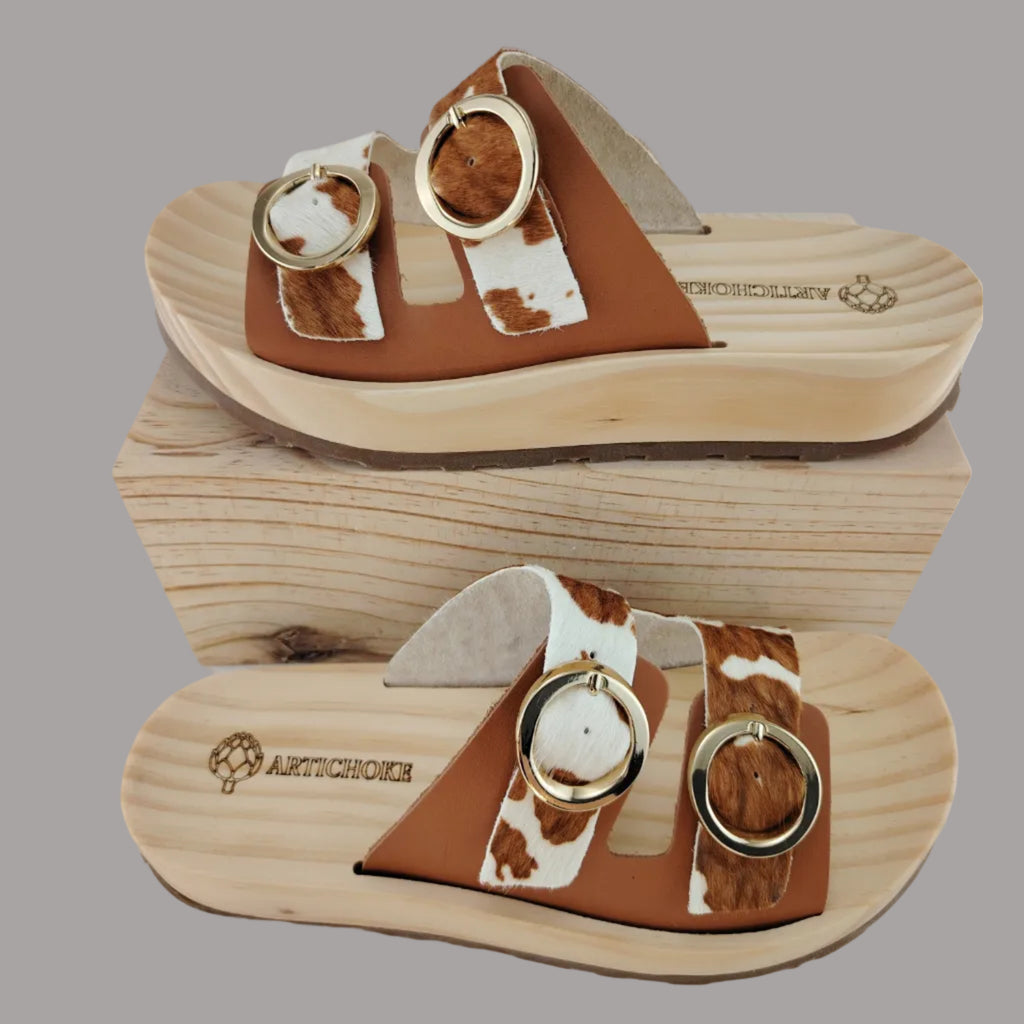 Sandalias tipo zueco de la marca Artichoke con base de madera, dos correas ajustables de cuero y piel sintética de estampado animal print de vaca, y hebillas redondas de metal dorado.