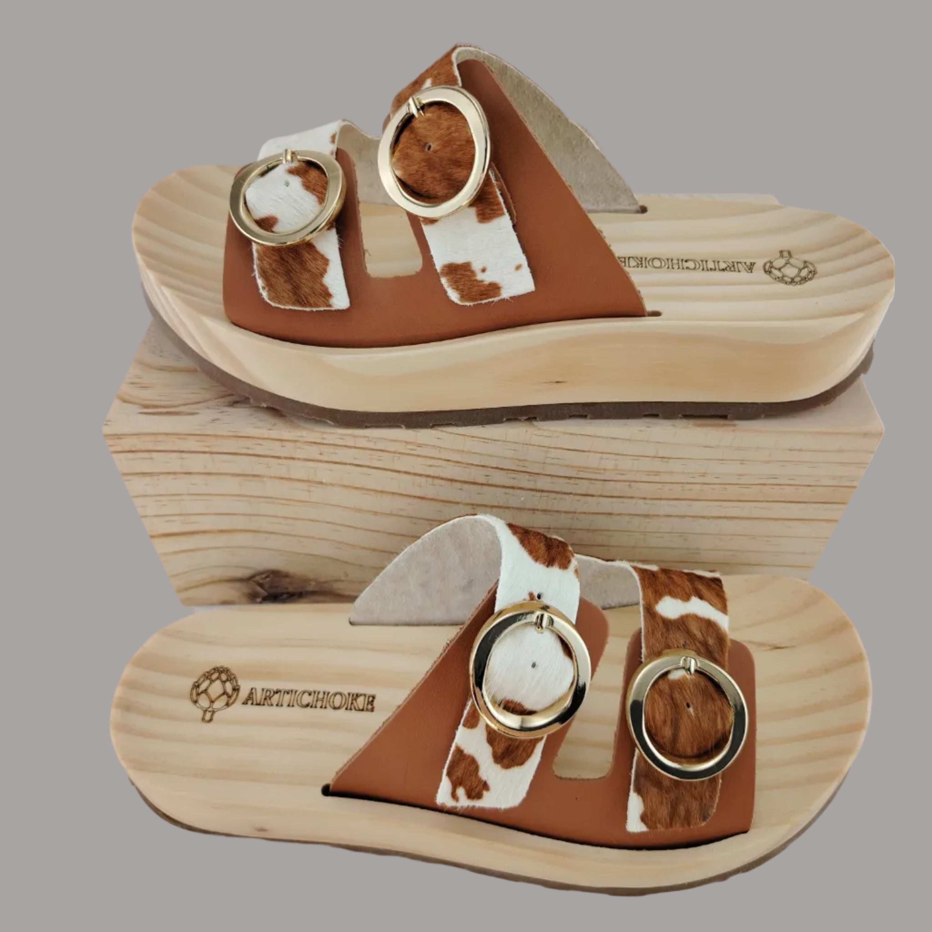 Sandalias tipo zueco de la marca Artichoke con base de madera, dos correas ajustables de cuero y piel sintética de estampado animal print de vaca, y hebillas redondas de metal dorado.