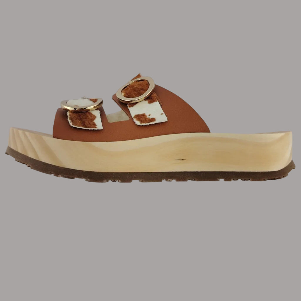 Sandalias tipo zueco de la marca Artichoke con base de madera, dos correas ajustables de cuero y piel sintética de estampado animal print de vaca, y hebillas redondas de metal dorado.