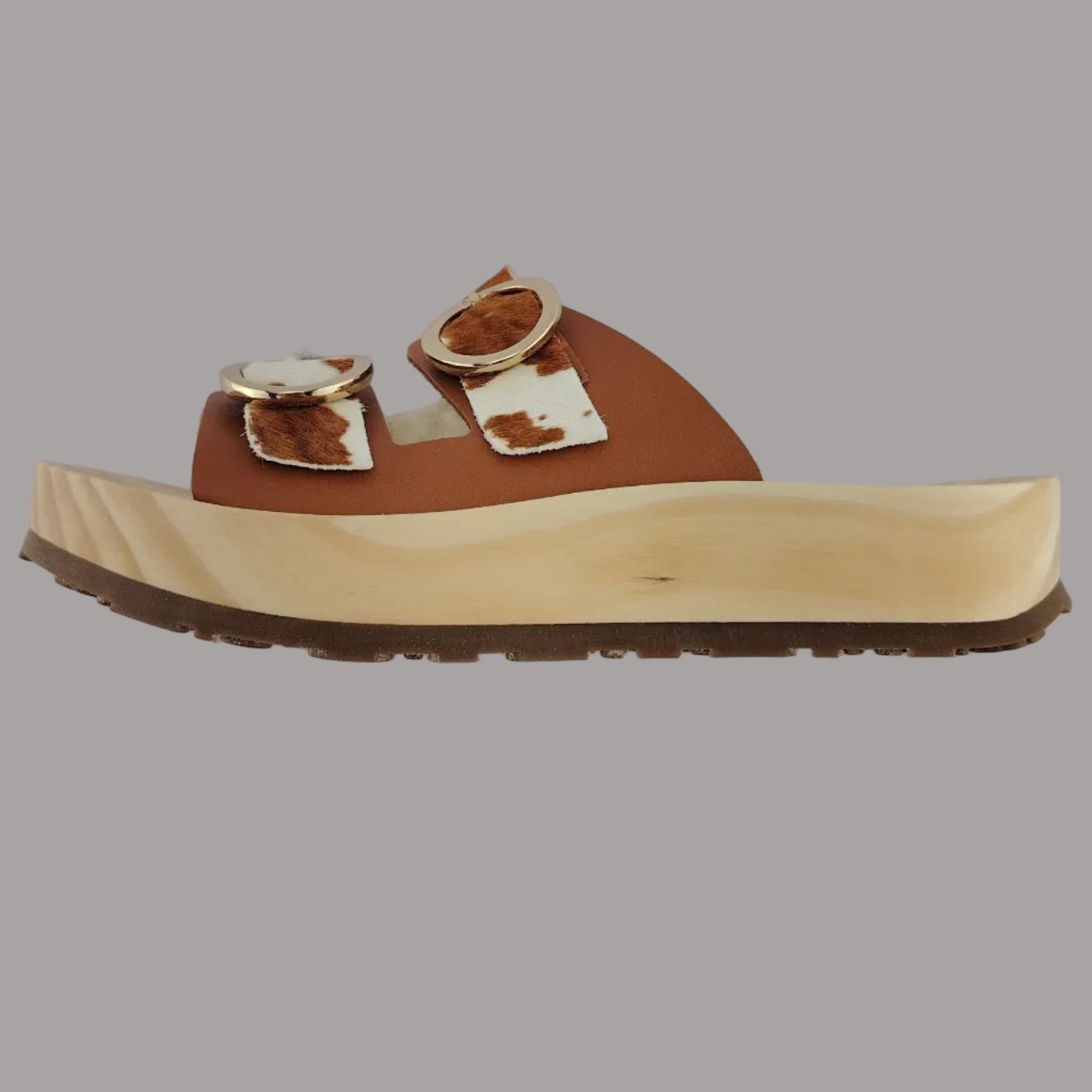 Sandalias tipo zueco de la marca Artichoke con base de madera, dos correas ajustables de cuero y piel sintética de estampado animal print de vaca, y hebillas redondas de metal dorado.