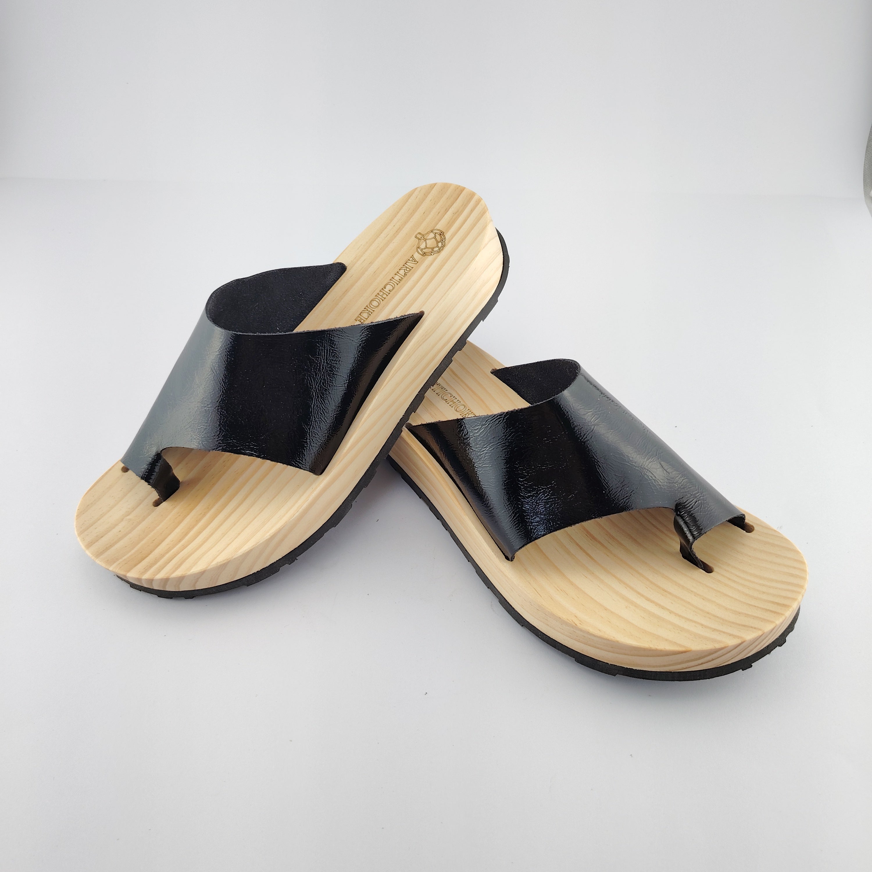Sandalias planas de madera natural artesanal con pala de piel negra brillante y sujeción de dedo, calzado de verano minimalista sobre fondo blanco.