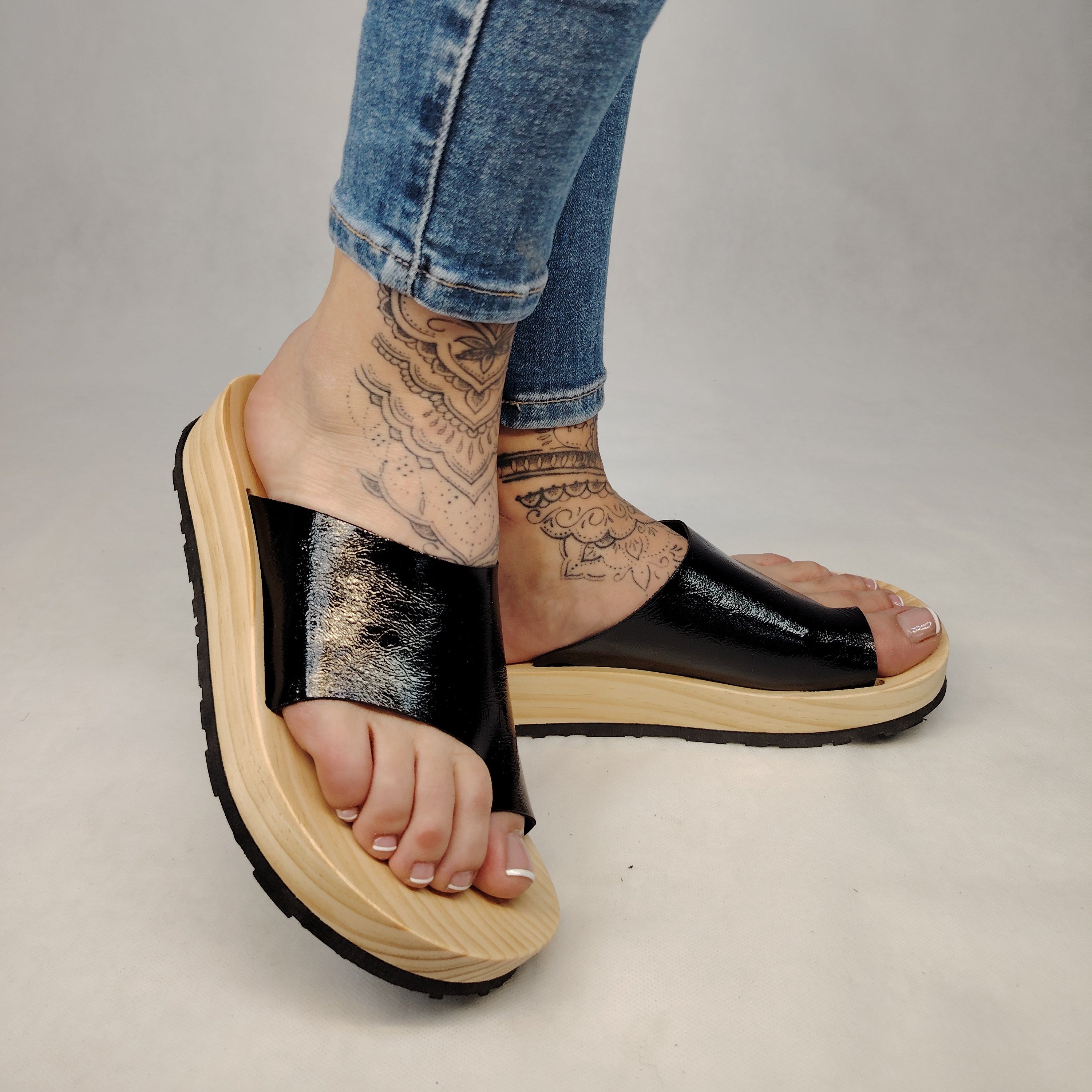 Sandalias planas de madera natural artesanal con pala de piel negra brillante y sujeción de dedo, calzado de verano minimalista sobre fondo blanco.