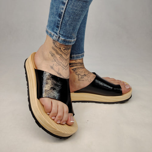 Sandalias planas de madera natural artesanal con pala de piel negra brillante y sujeción de dedo, calzado de verano minimalista sobre fondo blanco.