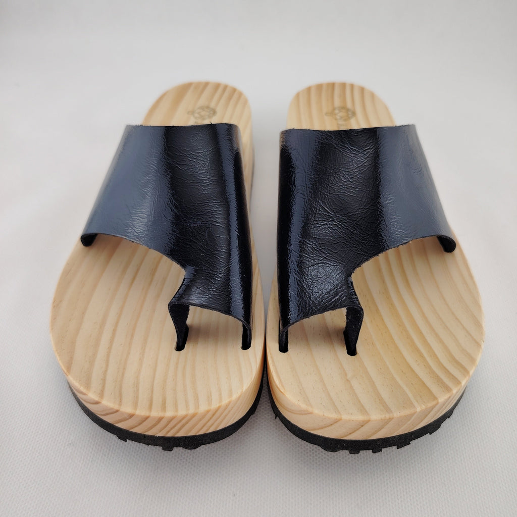 Sandalias planas de madera natural artesanal con pala de piel negra brillante y sujeción de dedo, calzado de verano minimalista sobre fondo blanco.