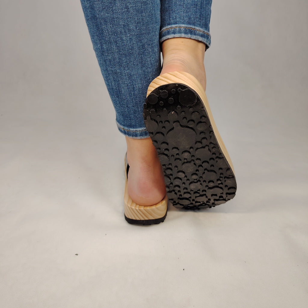 Sandalias planas de madera natural artesanal con pala de piel negra brillante y sujeción de dedo, calzado de verano minimalista sobre fondo blanco.