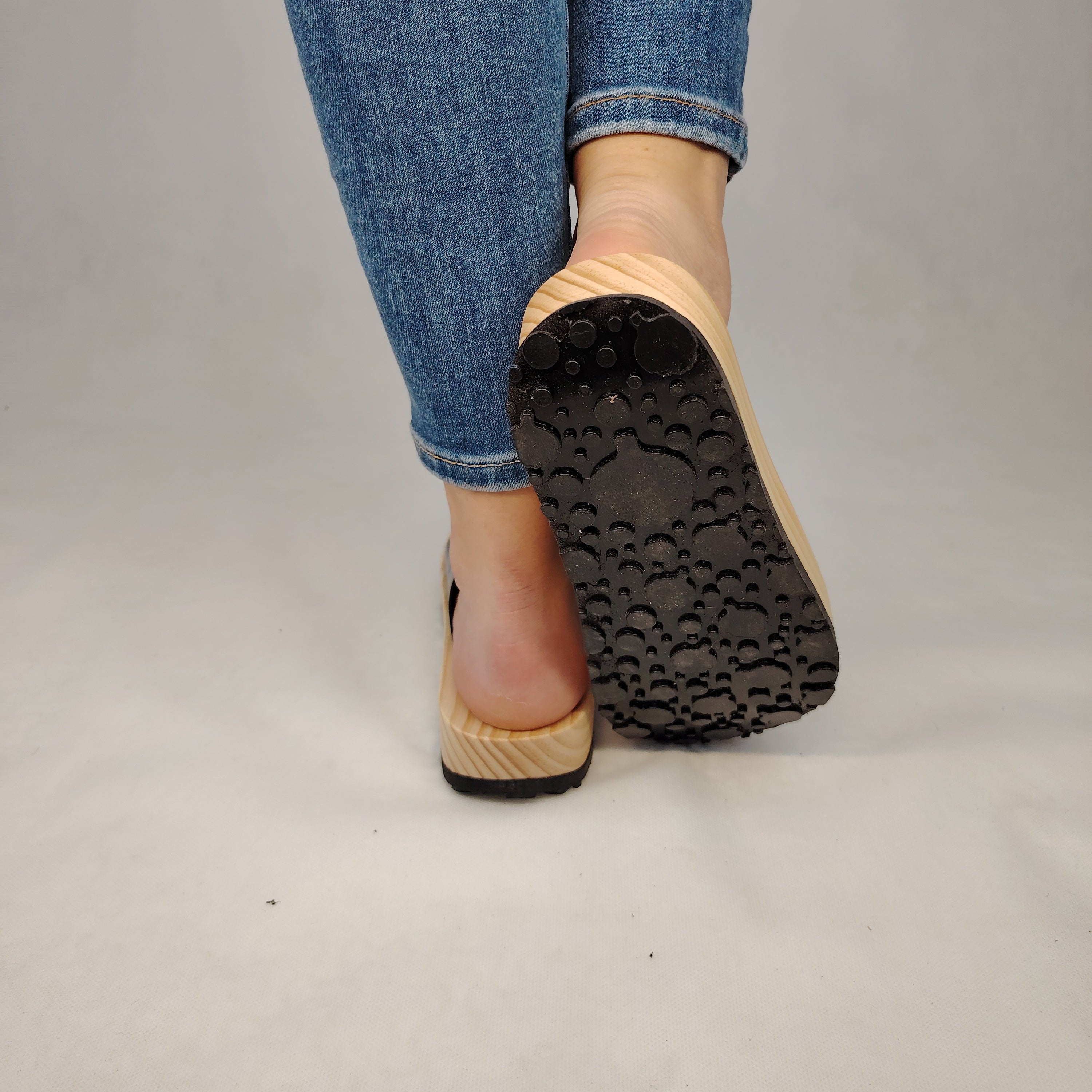 Sandalias planas de madera natural artesanal con pala de piel negra brillante y sujeción de dedo, calzado de verano minimalista sobre fondo blanco.