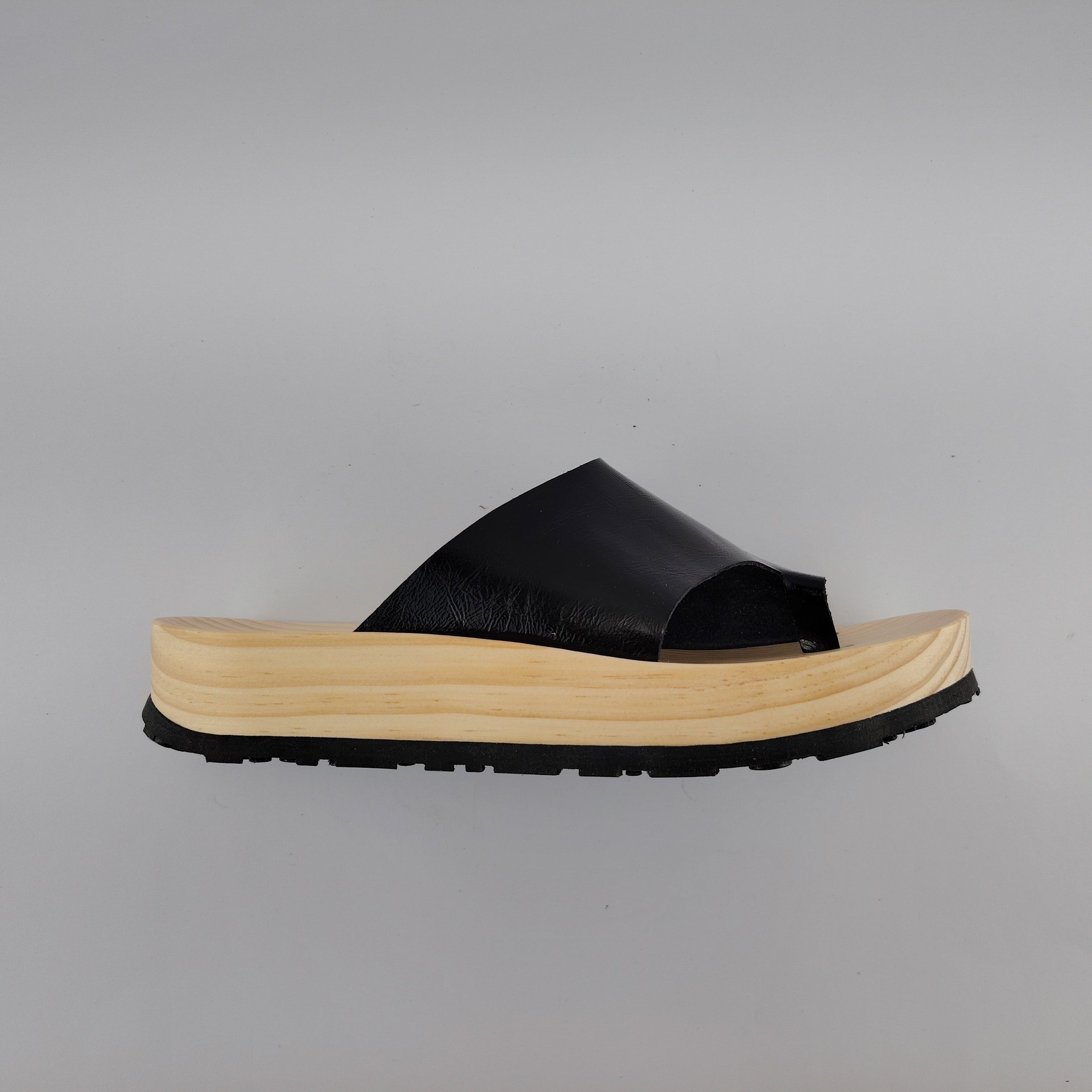 Sandalias planas de madera natural artesanal con pala de piel negra brillante y sujeción de dedo, calzado de verano minimalista sobre fondo blanco.