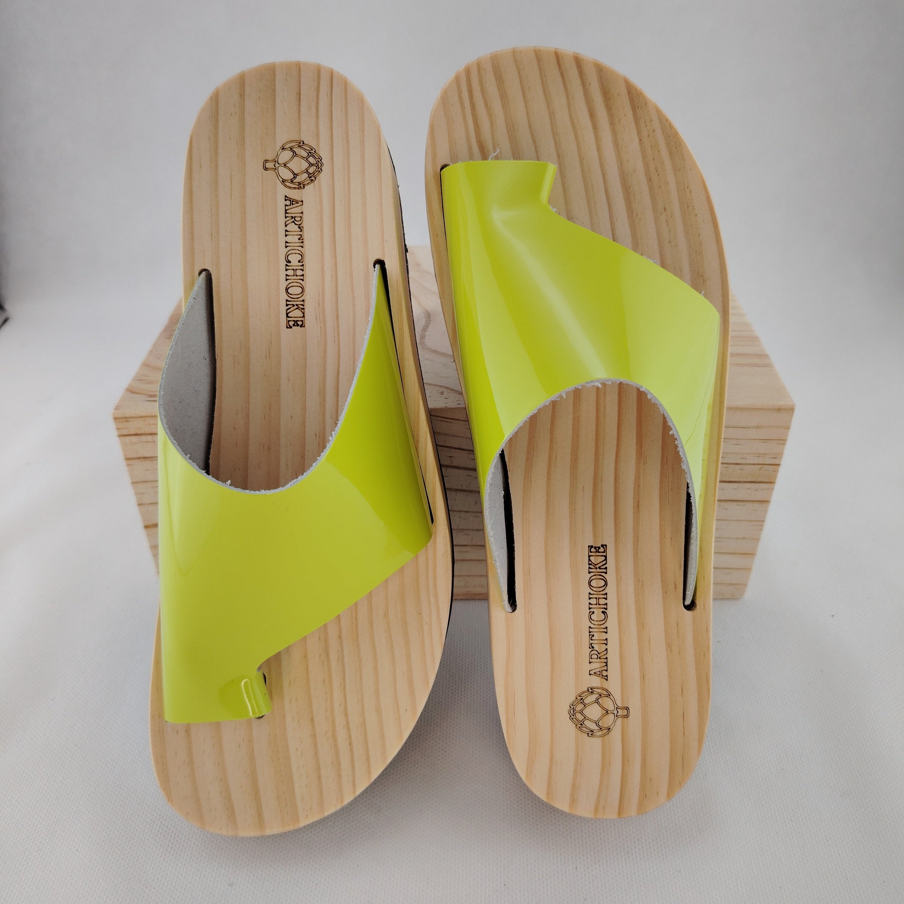 Sandalias planas de madera natural artesanal con pala de piel pistacho brillante y sujeción de dedo, calzado de verano minimalista sobre fondo blanco.