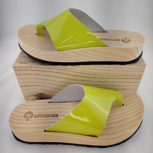 Sandalias planas de madera natural artesanal con pala de piel pistacho brillante y sujeción de dedo, calzado de verano minimalista sobre fondo blanco.