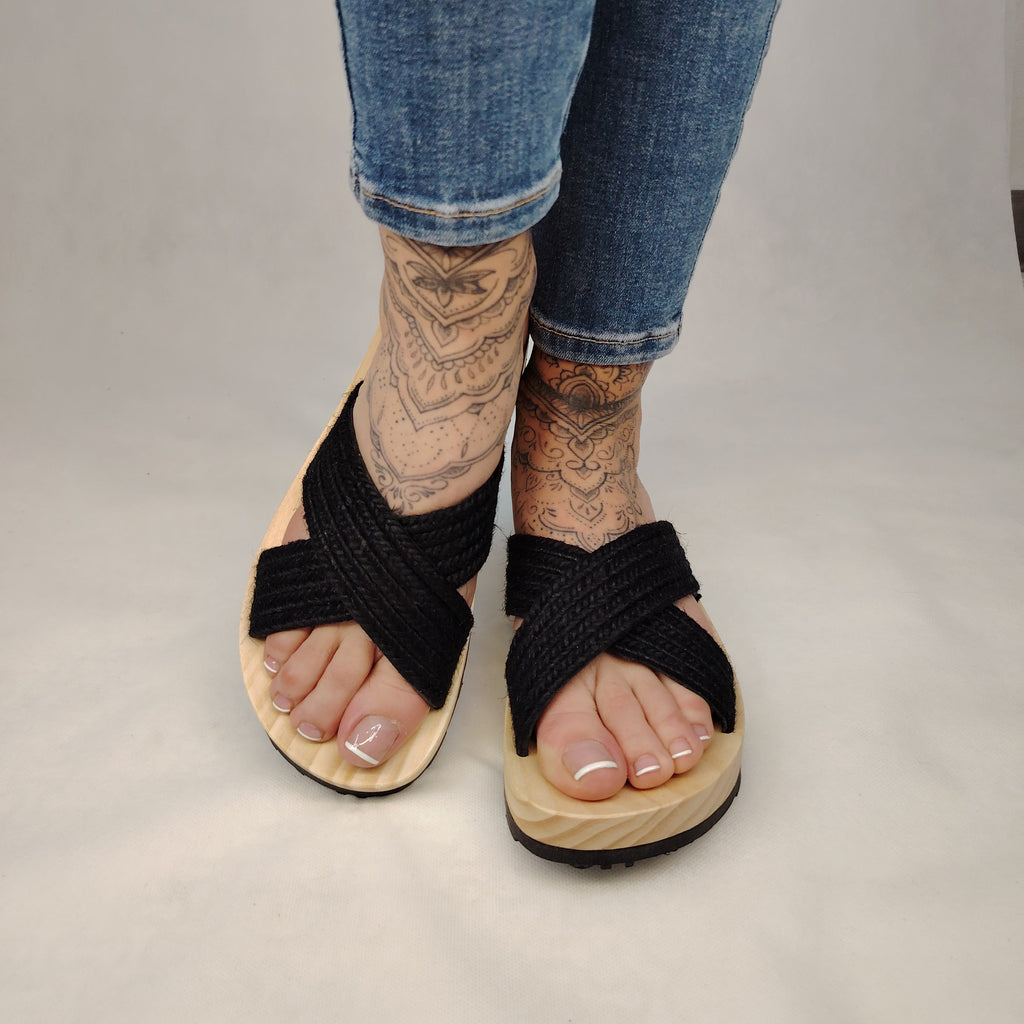 Sandalia plana de madera natural con tiras cruzadas de yute trenzada en color negro y diseño artesanal de verano.